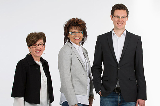 Drei Geschäftsleute stehen lächelnd in moderner Businesskleidung vor weißem Hintergrund.